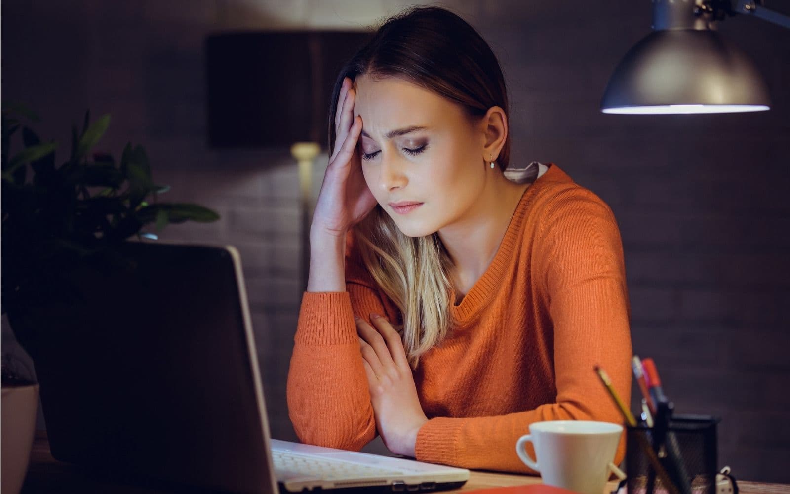 Γυναίκα κάθεται σε γραφείο με ανοιχτό laptop και στηρίζει το μέτωπό της στο χέρι.